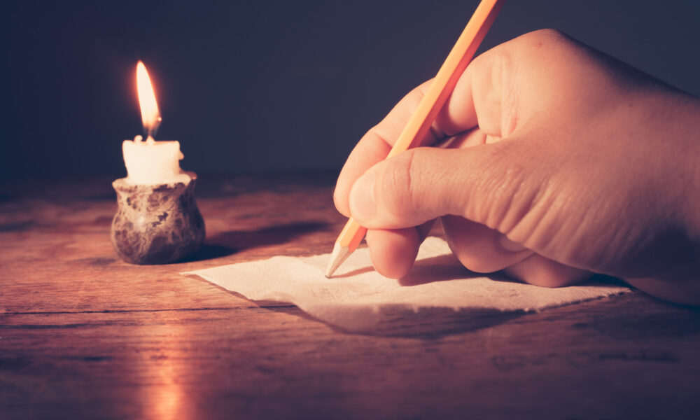 Hand schreibt mit Bleistift auf Papier bei Kerzenschein.