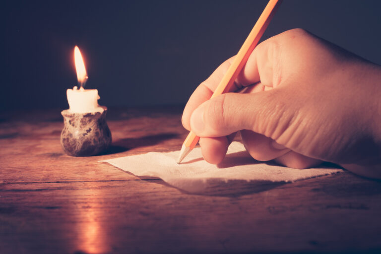 Hand schreibt mit Bleistift auf Papier bei Kerzenschein.