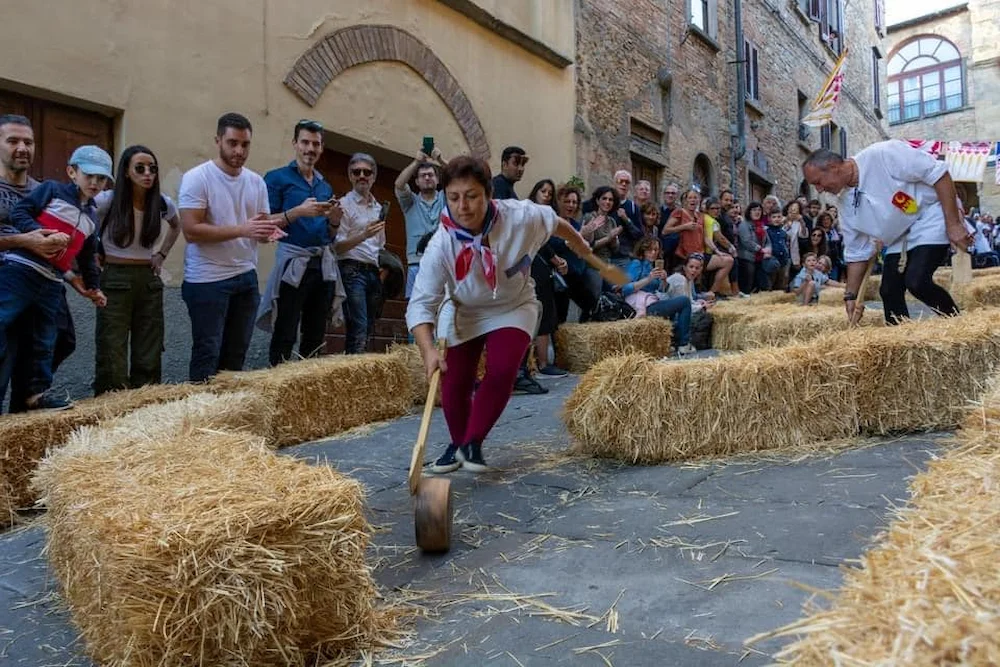 Toskana Wochenschau – Teilnehmerin beim Palio dei Caci in Volterra mit rollendem Käselaib zwischen Strohballen